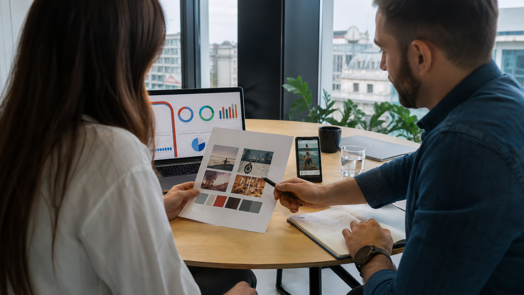 Doi specialiști de marketing analizează creative vizuale și dashboarduri pentru o campanie Demand Gen într-un birou modern